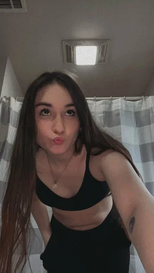 Woman with long dark hair in black sports bra and leggings, posing in bathroom with striped shower