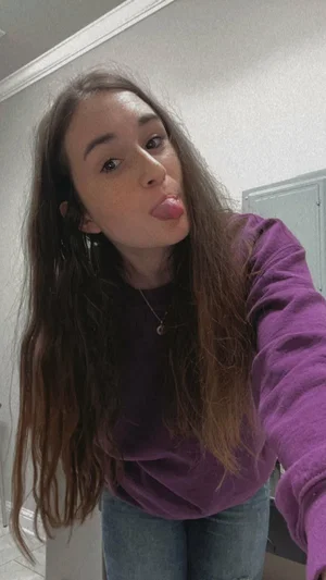 Woman with long brown hair in purple sweatshirt and jeans, making a playful face indoors.