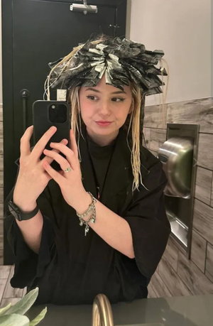 Woman with blonde hair in foils taking mirror selfie in bathroom.