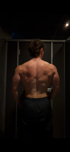 Shirtless muscular man standing back to camera in dark room wearing black pants.