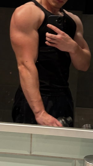 Muscular man in black tank top and shorts taking mirror selfie in bathroom.