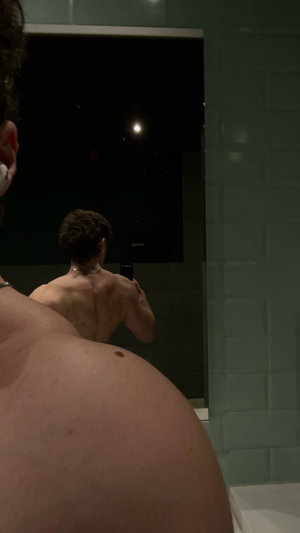 Shirtless man taking mirror selfie in bathroom, back visible, tiled walls.