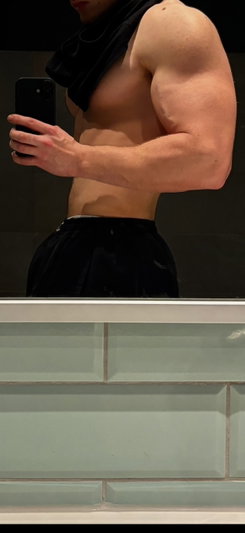 Shirtless muscular man in black shorts taking mirror selfie in bathroom.