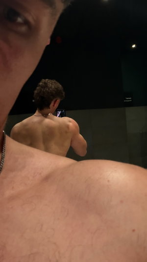 Shirtless man taking mirror selfie, showing back and shoulders in dimly lit room.