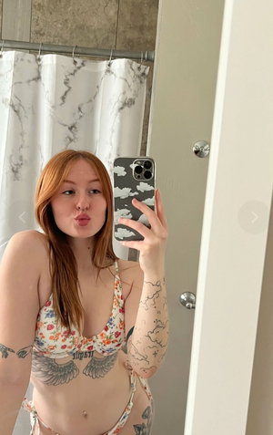 Red-haired woman in floral bikini taking mirror selfie in bathroom.