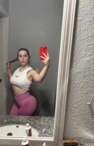 Woman in white top and pink leggings taking bathroom mirror selfie.