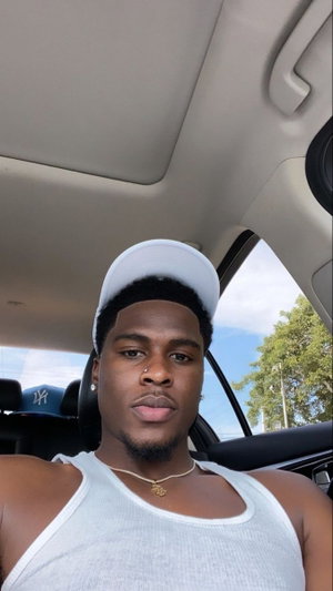Man in white tank top and cap sitting in car, looking at camera.