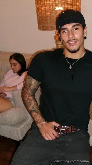 Man with tattoos and cap sitting on couch, woman in pink top in background.
