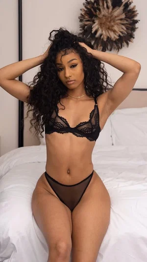 Woman with curly hair in black lace lingerie posing on a bed indoors.