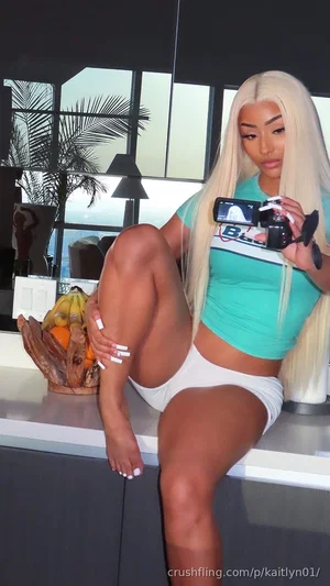 Woman with long blonde hair in teal crop top and white shorts, sitting on counter holding camera,