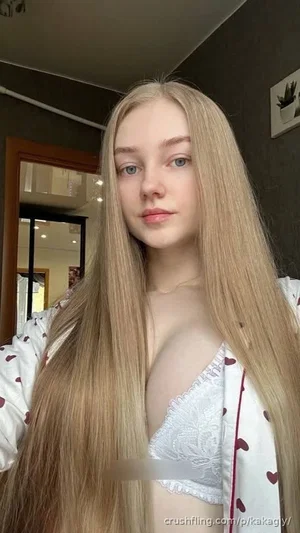 Blonde woman in white lace bra and heart-patterned robe, indoor selfie.