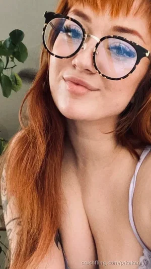 Woman with red hair and glasses, wearing a light-colored top, indoors with a plant in the