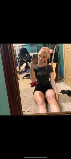 Woman with pink hair in black top and shorts takes mirror selfie in bedroom.