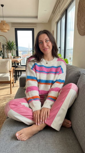 Woman in striped sweater and pink pants sitting on couch indoors.