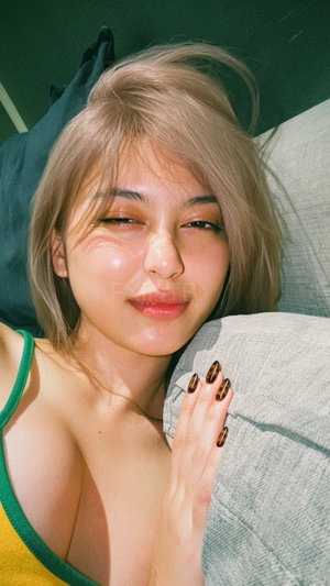 Woman with blonde hair in green and yellow top resting on gray pillow indoors.
