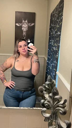 Woman with tattoos in gray tank top and jeans takes mirror selfie in bathroom.