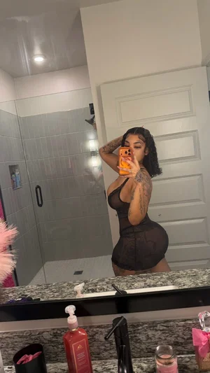 Woman in black sheer outfit with tattoos takes mirror selfie in bathroom.