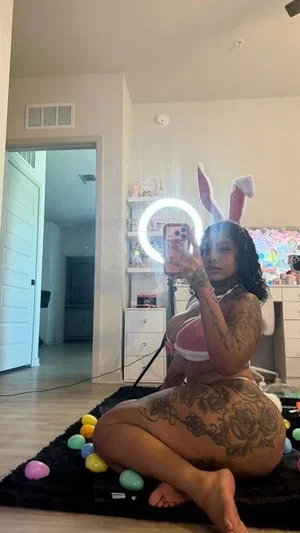 Woman with tattoos in bunny ears and pink lingerie, sitting on rug with colorful eggs indoors.