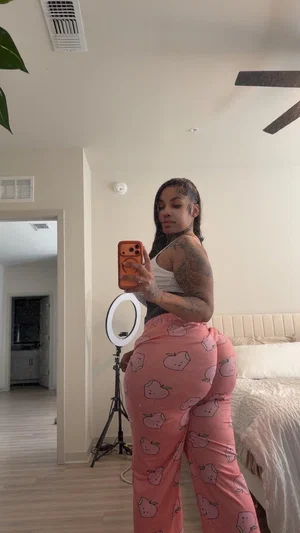 Woman with tattoos in pink patterned pants and white top taking mirror selfie in bedroom.