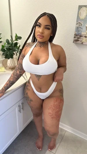 Woman with tattoos in white bikini posing in bathroom, long braids, indoor setting.