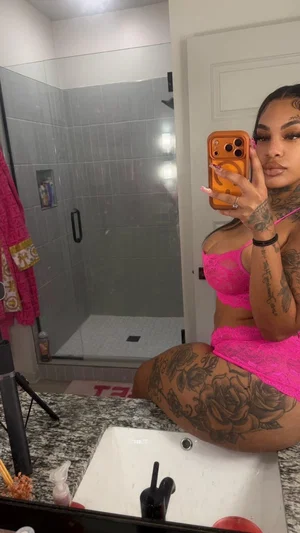 Woman with tattoos in pink lingerie sitting on bathroom sink, mirror selfie.