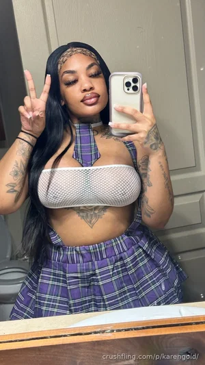 Woman with long black hair and tattoos wears mesh top and plaid skirt, taking mirror selfie in