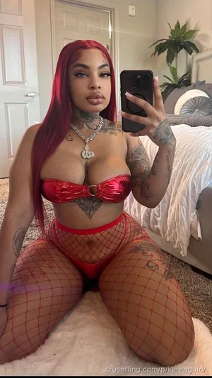 Woman with red hair and tattoos wears red lingerie and fishnet stockings, kneeling on a rug in a