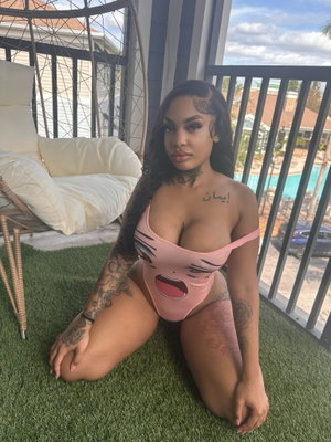 Woman in pink one-piece swimsuit with tattoos, kneeling on artificial grass on a balcony.