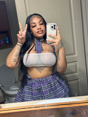 Woman in plaid skirt and mesh top taking mirror selfie in bathroom.