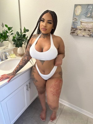 Woman with tattoos in white bikini posing in bathroom, braided hair, indoor setting.