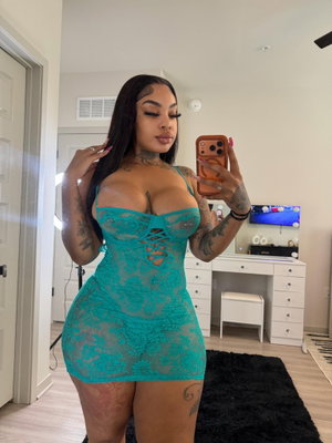 Woman in teal lace dress with tattoos takes mirror selfie in bedroom.