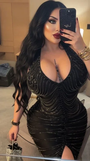 Woman with long black hair in a black sequined dress, holding phone for mirror selfie in bedroom.