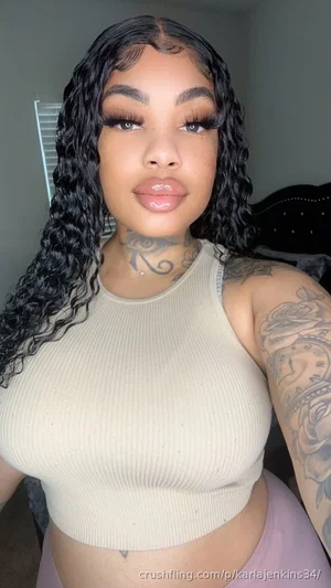 Woman with curly black hair, tattoos, wearing beige crop top and pink bottoms, indoors.