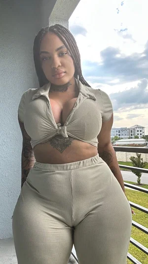 Woman with braids and tattoos wears beige ribbed crop top and pants, standing on balcony.