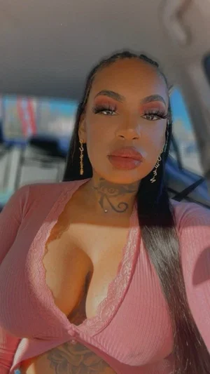 Woman with tattoos and long dark hair in a pink top, taking a selfie in a car.