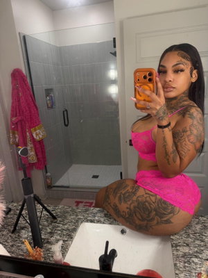 Woman with tattoos in pink lingerie sitting on bathroom counter, holding phone for mirror selfie.