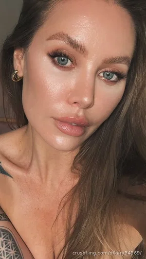 Woman with long brown hair and blue eyes, wearing makeup, showing chest tattoo, indoors.