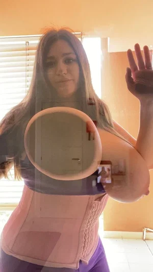 Woman with long hair wearing pink corset and purple pants, holding mirror, waving indoors.