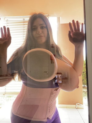 Woman with long hair wearing pink corset and purple pants, posing by glass door with hands raised.