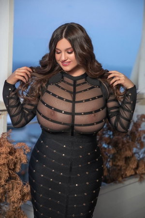 Woman in black sheer dress with studded details, posing indoors near plants.