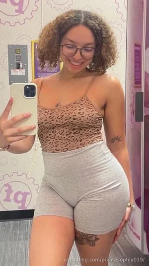 Woman with curly hair in leopard print top and gray shorts, smiling in mirror selfie.