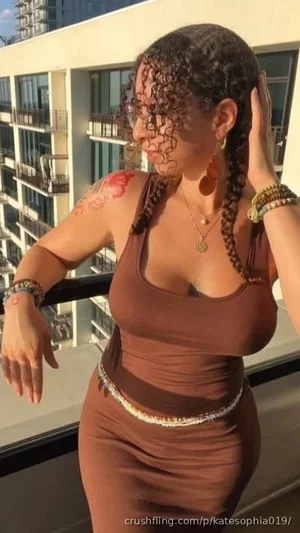 Woman with curly hair in braids, wearing brown tank top and skirt, standing on balcony with