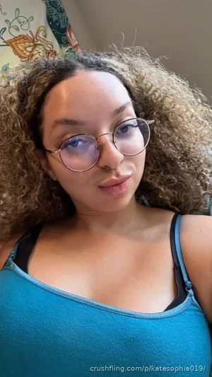 Woman with curly hair and glasses wearing a blue tank top indoors.