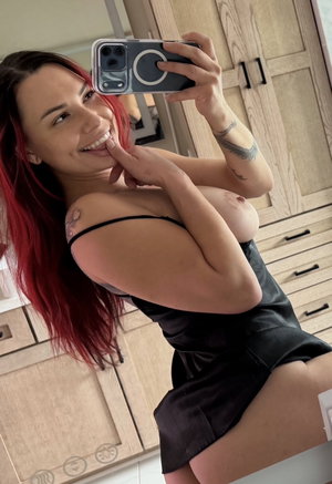 Red-haired woman in black dress taking mirror selfie indoors.