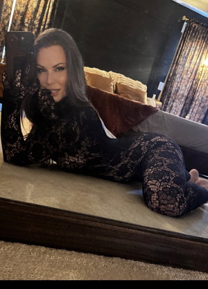Woman in black lace bodysuit taking mirror selfie on bed.