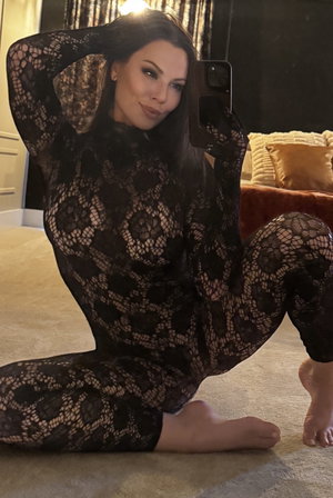 Curvy brunette in black lace bodysuit taking selfie on bedroom floor.