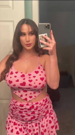 Woman in pink heart-patterned crop top and skirt takes mirror selfie indoors.