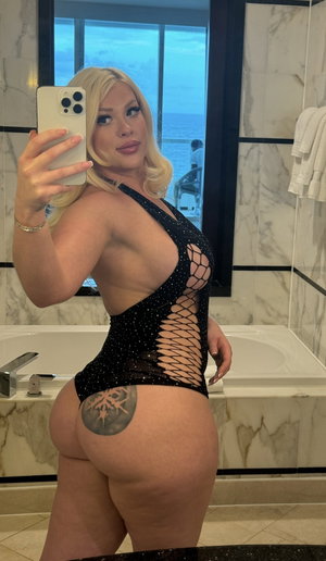 Blonde woman in black mesh bodysuit with tattoo, taking mirror selfie in marble bathroom.