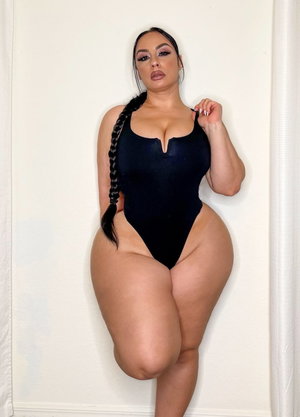 Woman in black bodysuit posing against white wall, long braided hair, curvy figure.