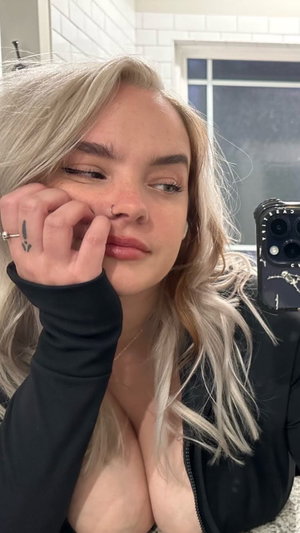 Blonde woman in black top, posing in bathroom with hand near face.